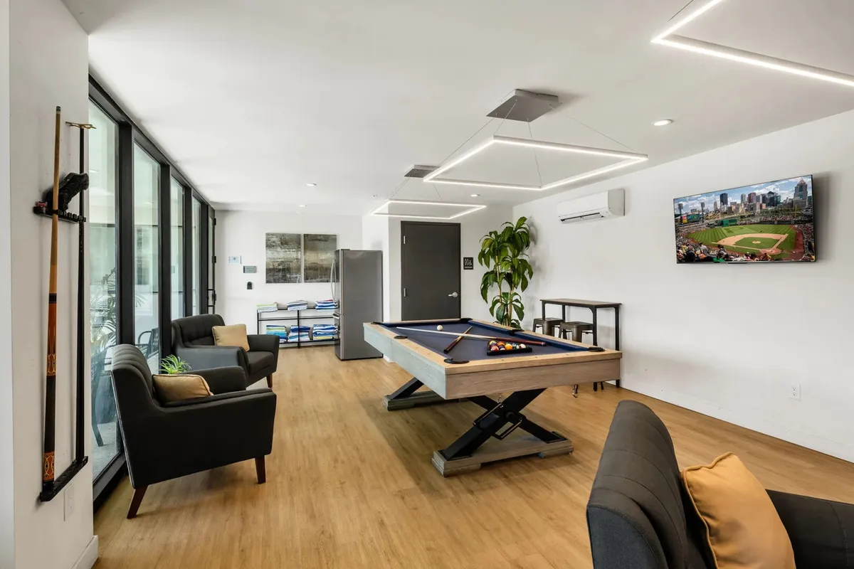 A stylish interior clubhouse featuring a pool table, a large TV screen, comfortable seating, and green plants. The space is bright and welcoming, designed for casual gatherings, social games, or watching sports with friends.