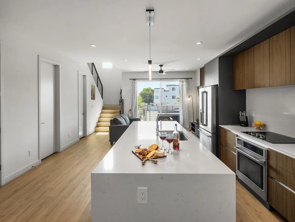 A wide angle captures the kitchen island, modern appliances, a comfortable seating area, and a staircase. Natural light pours in through large windows, giving the space a welcoming, contemporary atmosphere. The kitchen and living area flow together, ideal for gatherings and daily living.