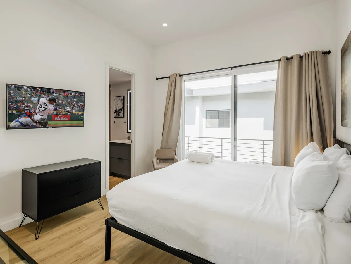 This image displays a serene bedroom with a king bed dressed in white linens, a wall-mounted television, a modern dresser, and a large window with curtains that lets in gentle natural light. The room feels spacious, serene, and ideal for unwinding.
