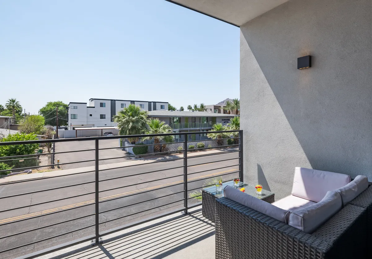 This image displays a private balcony with comfortable seating, palm trees, and a street view. The inviting setup is ideal for enjoying coffee in the morning or unwinding at sunset, echoing the described transition from kitchen to outdoor relaxation.