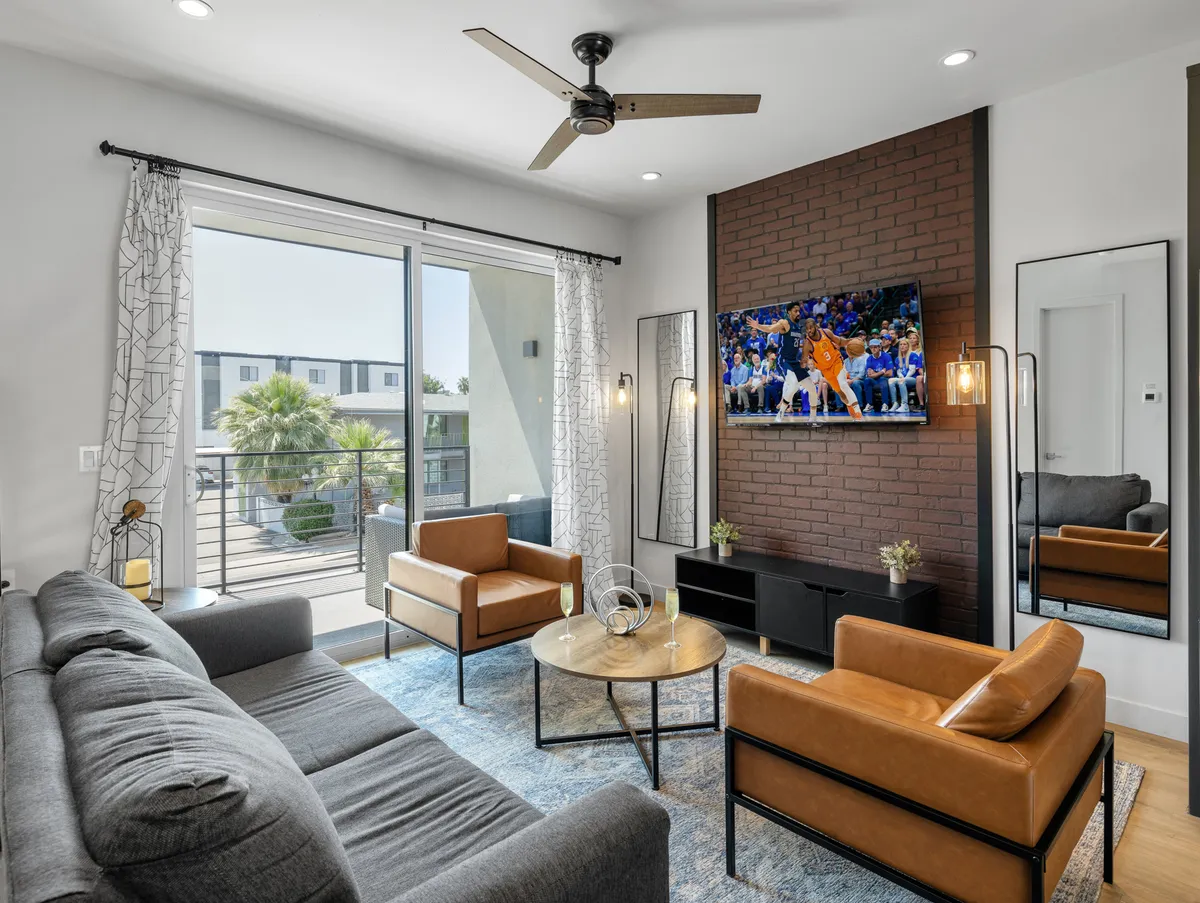 The image showcases a chic, modern living room filled with natural light from a large window. A plush sofa and stylish armchairs surround a coffee table, positioned in front of a flat-screen television displaying a live sports game. The inviting setup suggests a space perfect for socializing, relaxing, or catching the big game. Outside the window, you catch a glimpse of Scottsdale’s vibrant cityscape.