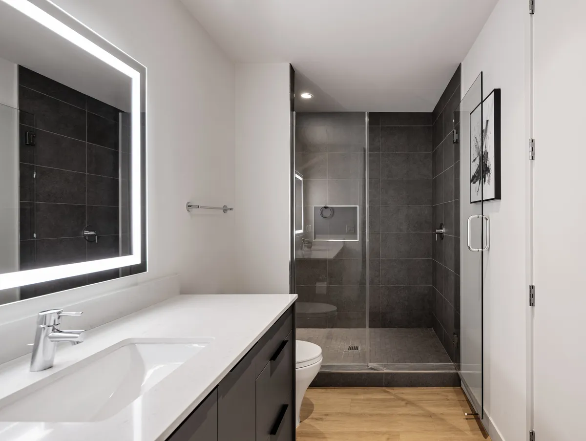 A contemporary en-suite bathroom is featured, complete with an illuminated mirror, minimalist sink, and a glass shower enclosure. The design is sleek and spa-like, emphasizing relaxation and modern comfort.