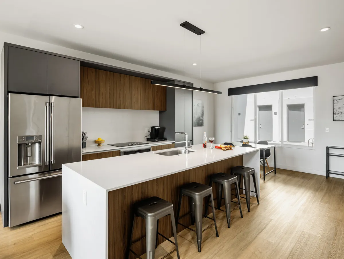 A bright, contemporary kitchen with a large island, stainless steel appliances, wood cabinetry, and abundant natural light streaming through the windows. The setting feels inviting for both cooking and socializing, with a layout that suggests functional ease and thoughtful amenities.