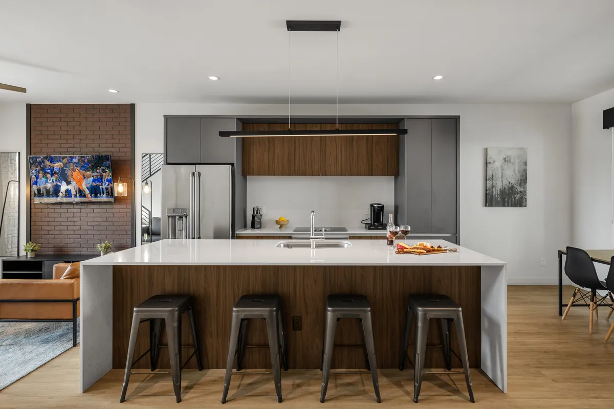 This image showcases an open-plan living space that includes a large kitchen island with seating, stainless steel appliances, wooden cabinetry, and a smart TV mounted on the wall. The layout is designed for both cooking and socializing, with the kitchen and living room visually connected, hinting at the comfort and multi-functionality described in the section.