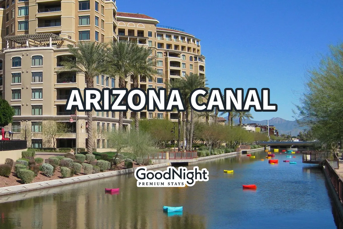 A lively, sun-dappled stretch of the Arizona Canal, flanked by palm trees and colorful boats, with people strolling along the waterway and modern residential buildings in the background. The scene feels vibrant and inviting, suggesting a community hub where locals and visitors enjoy outdoor activities together.